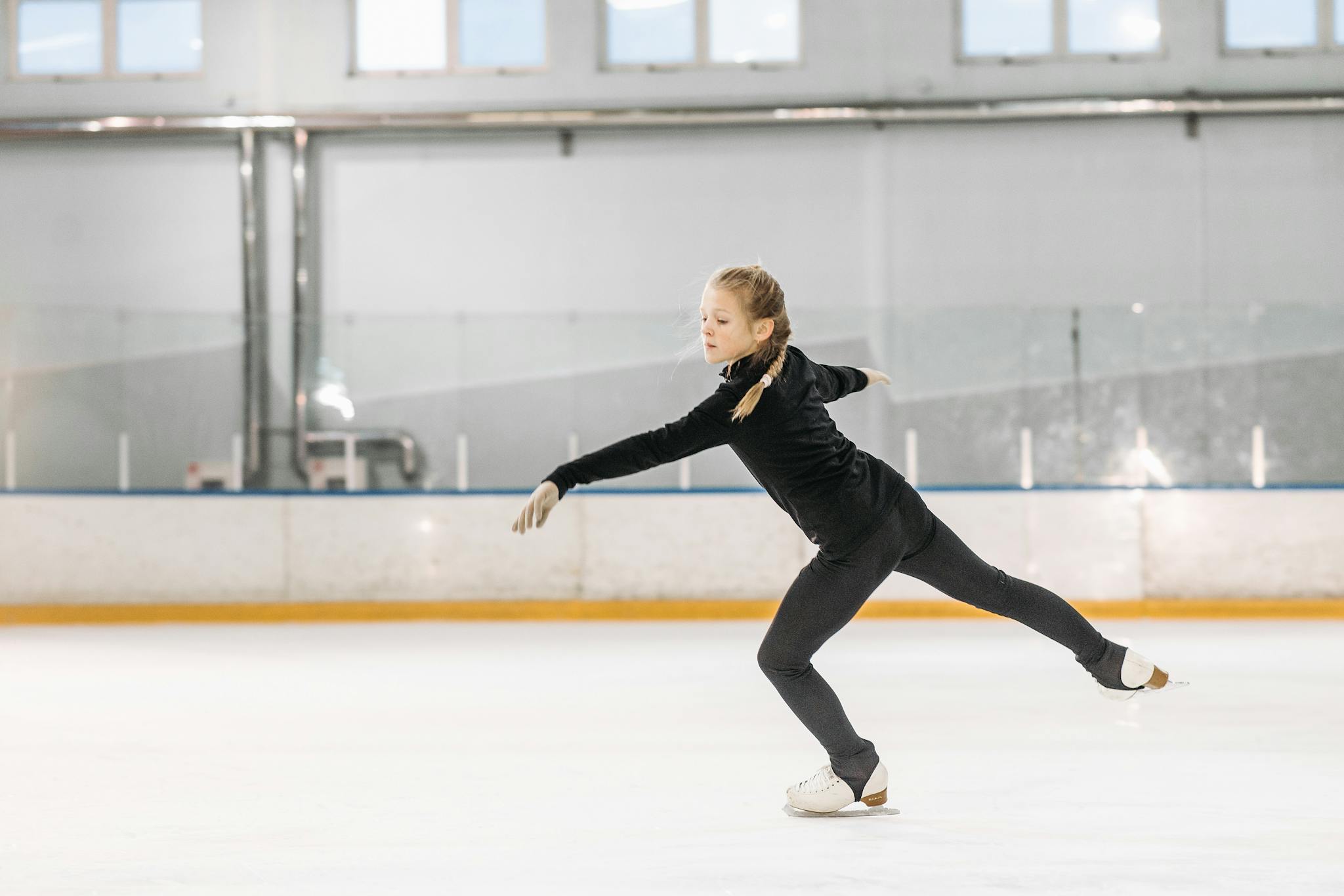 A young girl practicing figure skating moves at an indoor ice rink during the day.